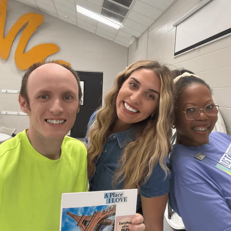Three smiling people pose together in a room with white walls and a yellow decoration, while the person in the center holds a card featuring the Eiffel Tower.