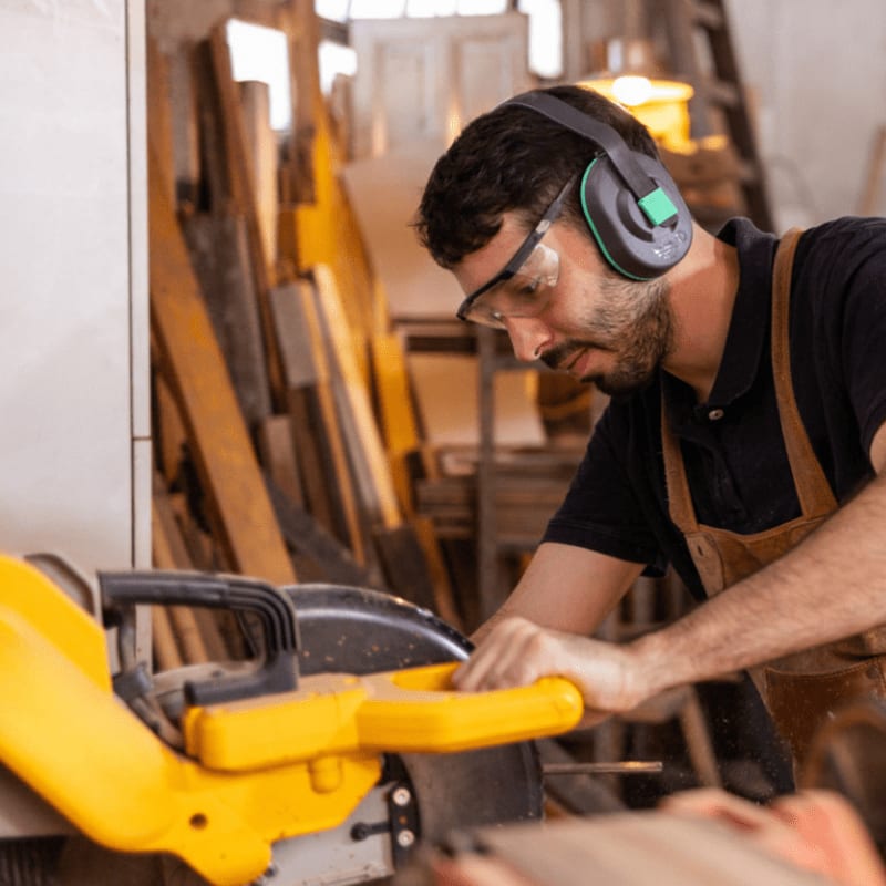 A man wearing safety gear and an apron works with a yellow power saw in a woodworking shop filled with lumber.