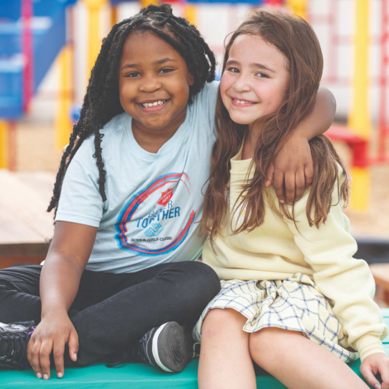 Two smiling young girls sit closely together on a green bench with a colorful playground blurred in the background.