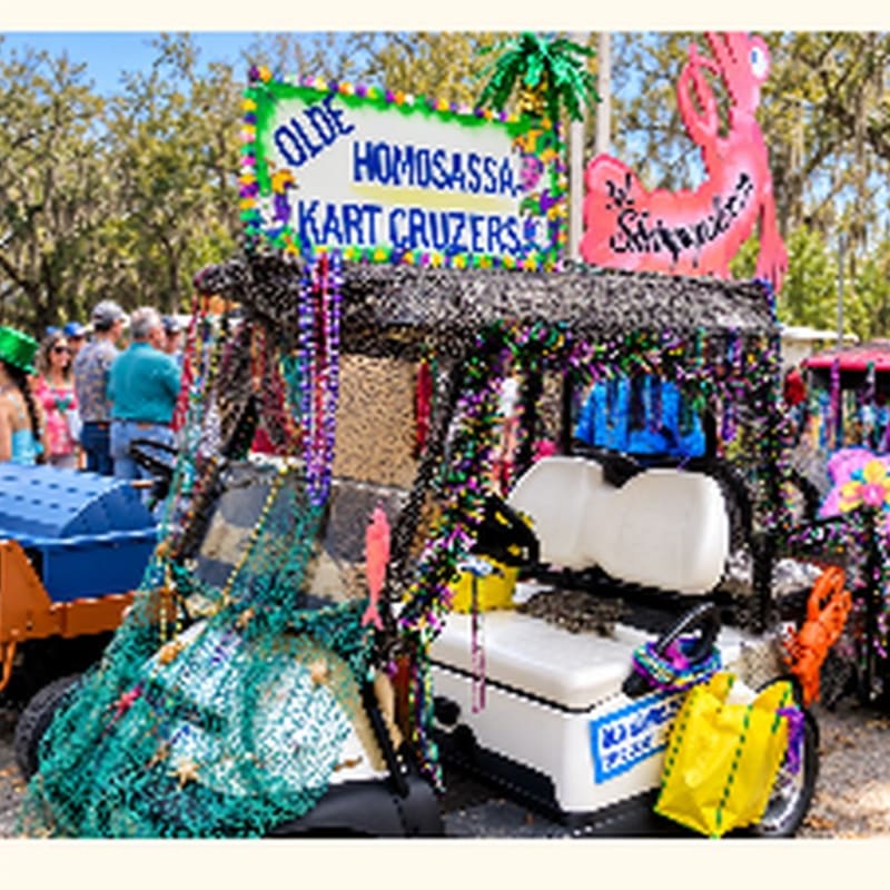 A decorated golf cart with a "Olde Homosassa Kart Cruzers" sign and colorful beads sits in the foreground against a background of trees and people.