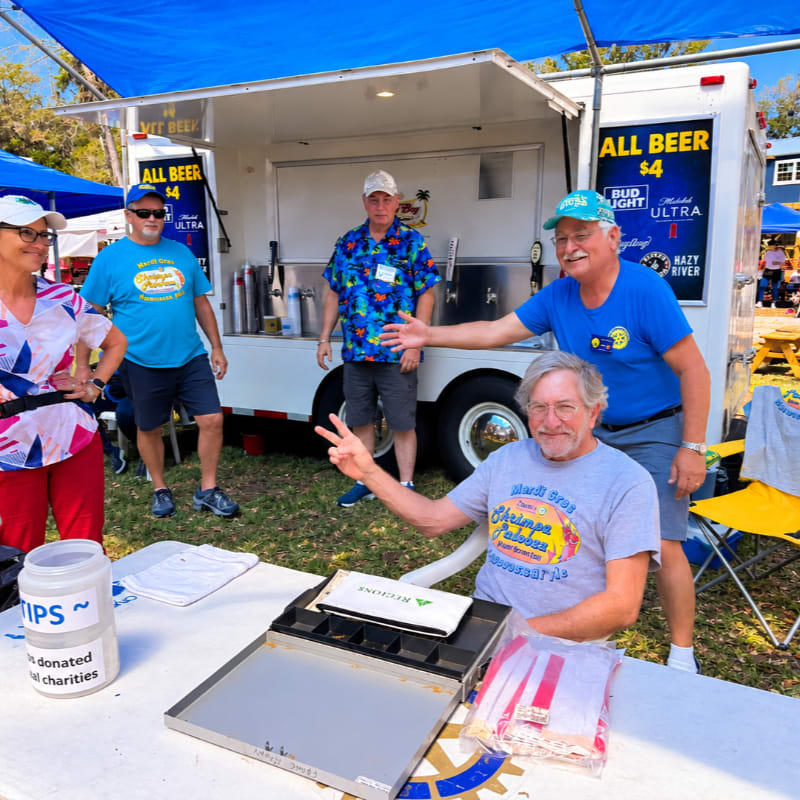 Three men and a woman stand near a white beer trailer under a blue canopy in an outdoor grassy area.