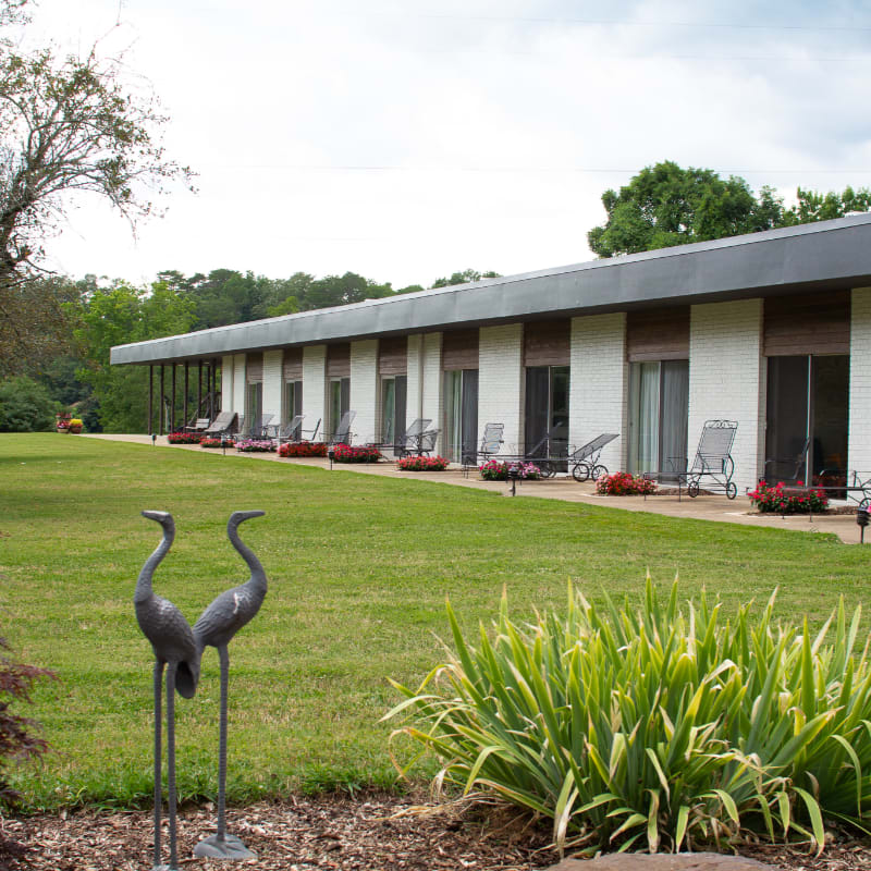 Two metal crane sculptures and green foliage stand in the foreground of a grassy lawn leading to a long, single-story white motel under a cloudy sky.