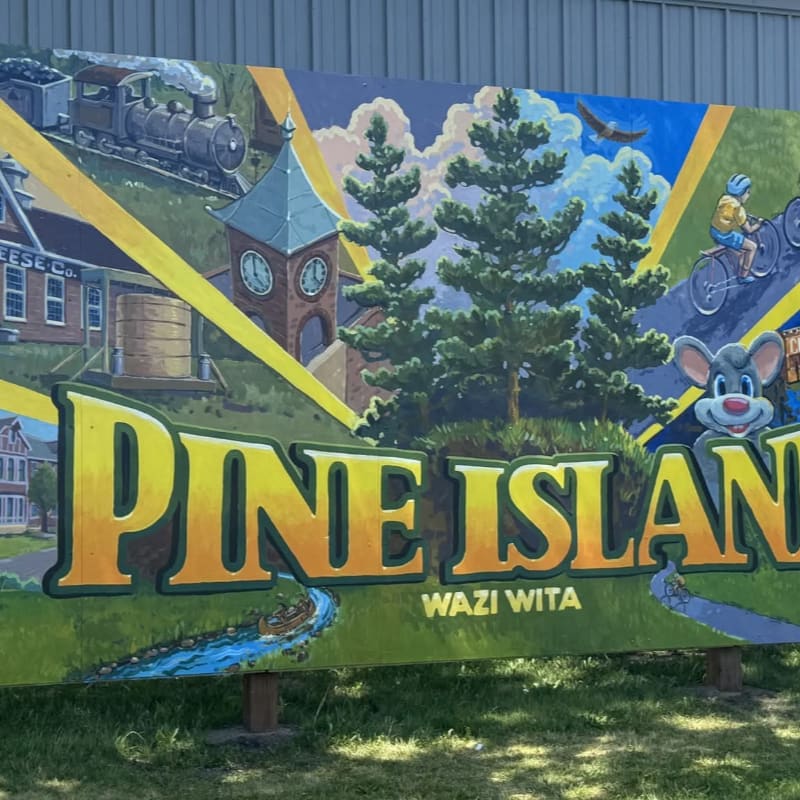 A colorful "Pine Island" sign stands in a grassy area, featuring illustrations of a train, a clock tower, pine trees, and a cyclist against a blue sky.