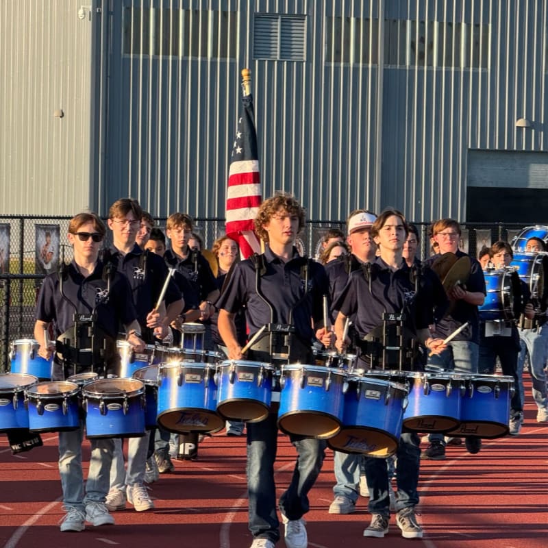 A marching band with blue drums and uniforms stands in front of a building with an American flag.
