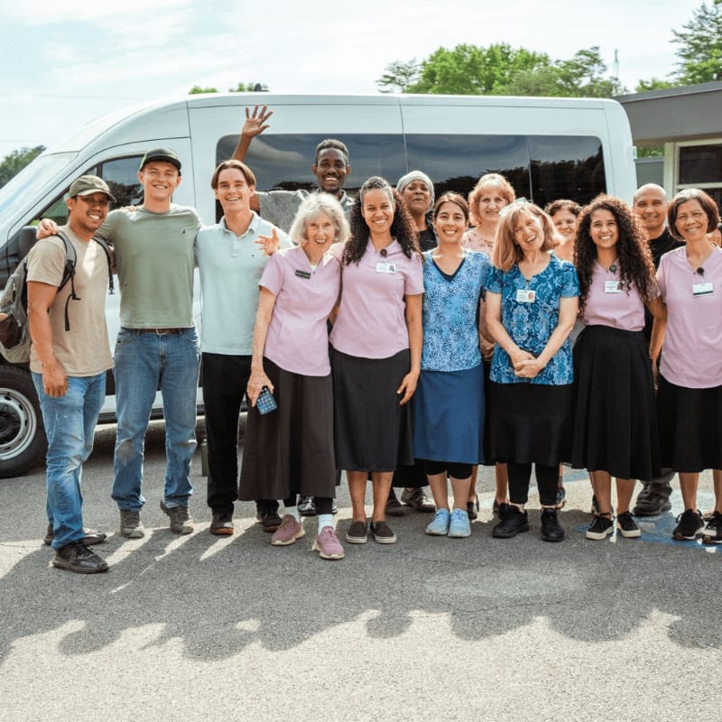 A diverse group of people poses for a photo in the foreground, standing in front of a white van and green trees in the background.