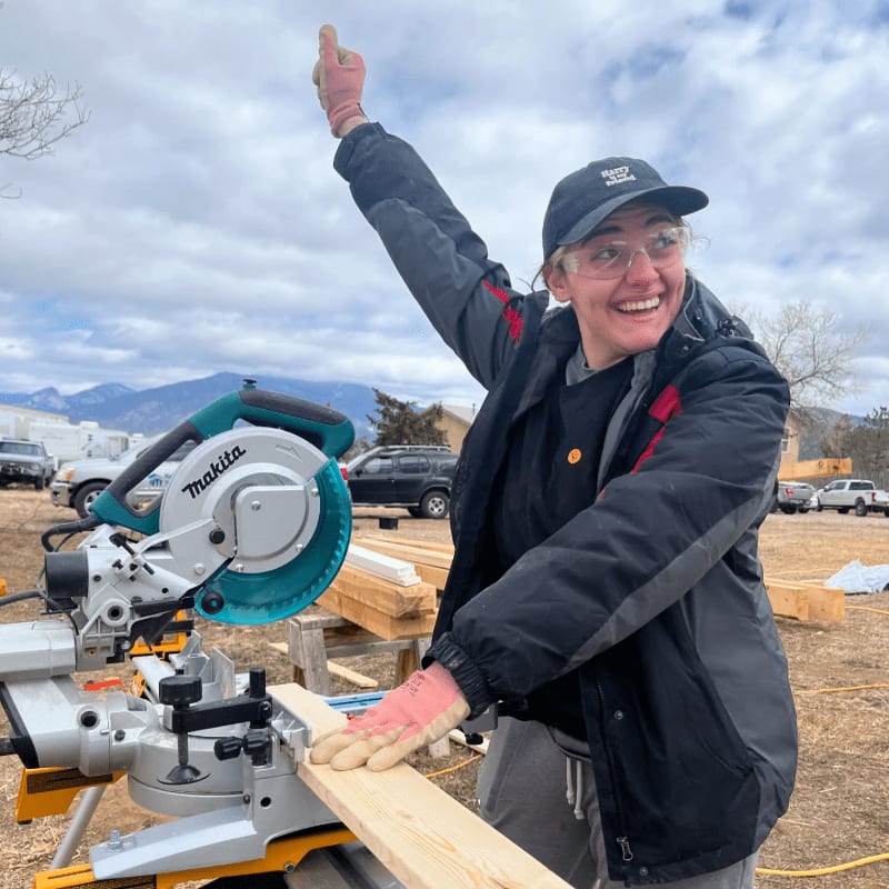 A smiling woman in a black jacket and safety gear gives a thumbs-up while operating a miter saw in an outdoor construction area with mountains in the distance.