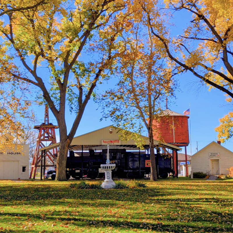A black train sits under a shelter in a grassy yard with autumn trees, a red water tower, and a small white building under a clear blue sky.