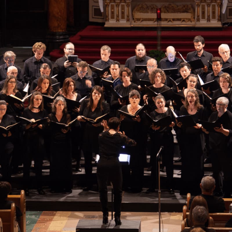 A choir of singers dressed in black performs on a stage, led by a conductor in the foreground against a backdrop of a grand hall with red carpeting and ornate architecture.