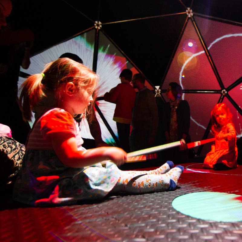 A young girl sits in the foreground of a geodesic dome featuring colorful projections and other people in the background.