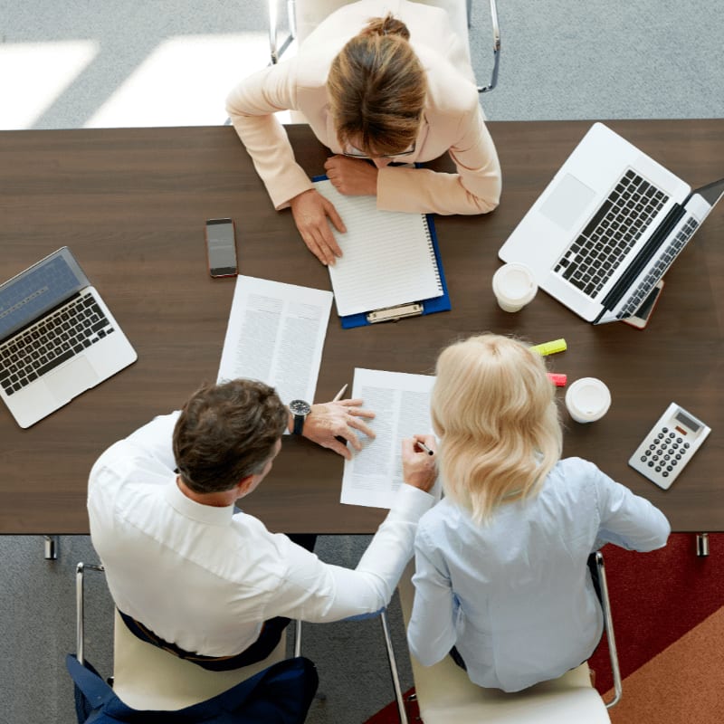 Three professionals sit around a dark wooden table covered with laptops, documents, and calculators in a bright office setting.