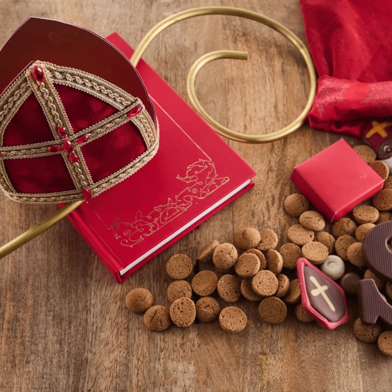 A red miter, a red book, and a golden staff sit beside a pile of small cookies and chocolates on a wooden surface.