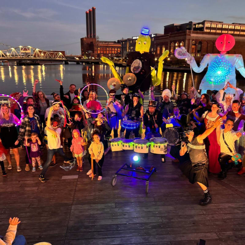 A diverse group of people in colorful costumes and large puppets pose on a wooden pier with a city skyline and river in the background.