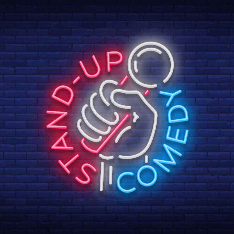 A neon sign of a hand holding a microphone with the words "STAND-UP COMEDY" glows against a dark blue brick wall.