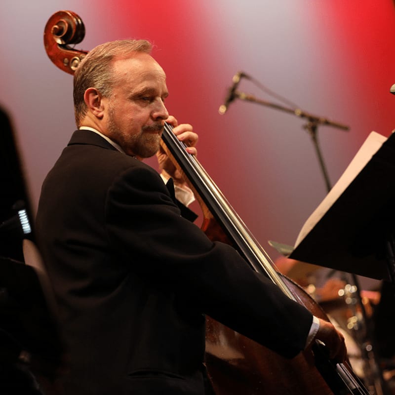 A man in a black suit plays a double bass in the foreground against a blurred red and white background.