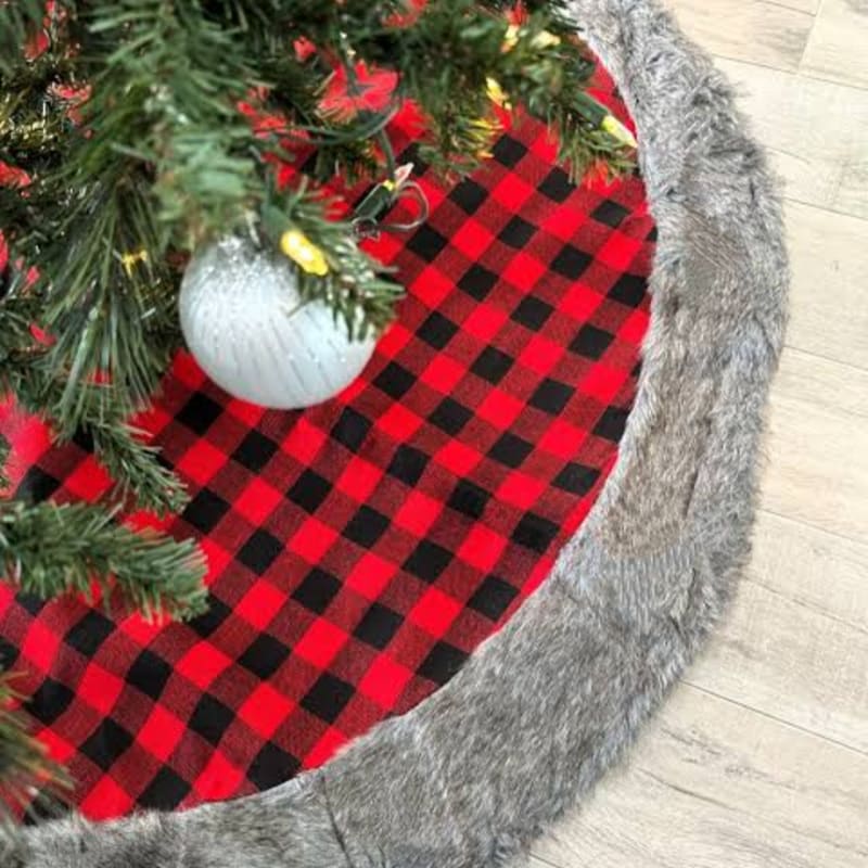A Christmas tree with a silver ornament sits atop a red and black buffalo plaid tree skirt with a grey faux-fur border on a light wood floor.