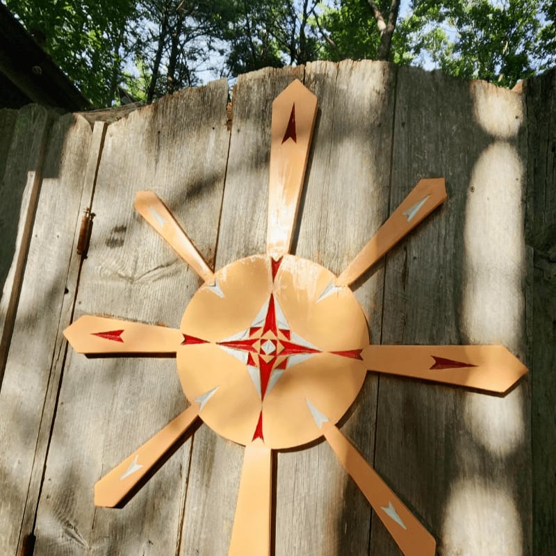 A tan, star-shaped wooden compass rose is mounted on a weathered wooden fence with green trees in the background.
