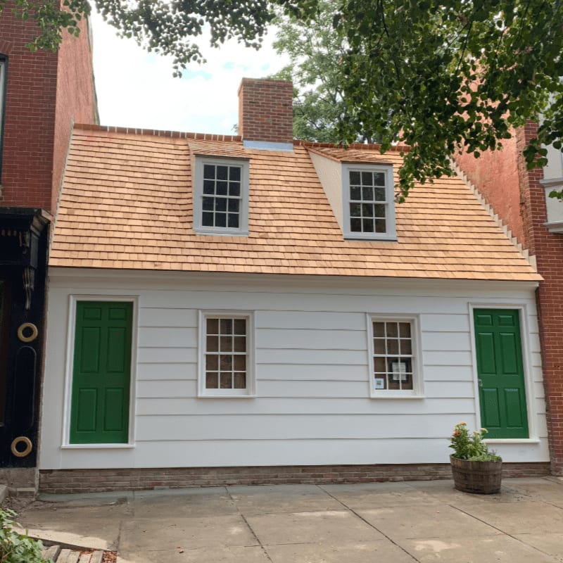 A small white house with a cedar shake roof and green doors stands in a paved courtyard, flanked by brick buildings and overhanging green leaves.