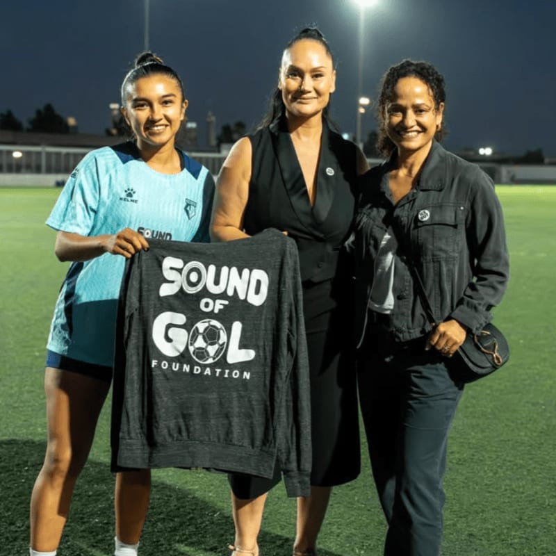 Three women stand on a green soccer field at night, with two of them holding up a dark grey "Sound of Gol Foundation" sweatshirt.