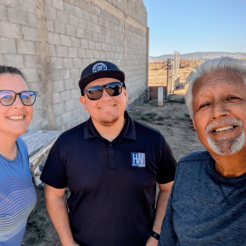 Three smiling people stand in the foreground against a background of a concrete block wall and a distant hilly landscape.