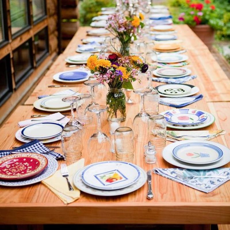 A long wooden table is set with colorful plates, glassware, and floral centerpieces, set against a background of a rustic building and garden greenery.