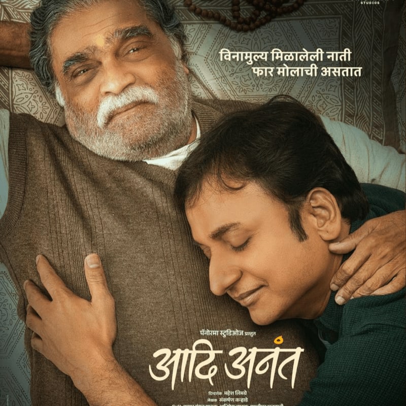 A younger man rests his head on the chest of an older, bearded man in the foreground, set against a patterned fabric background with Marathi text.