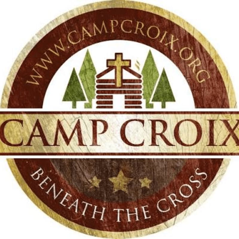A circular logo features a log cabin with a cross and pine trees in the center, framed by a brown border containing a website URL and the phrase "BENEATH THE CROSS," with a central banner reading "CAMP CROIX."