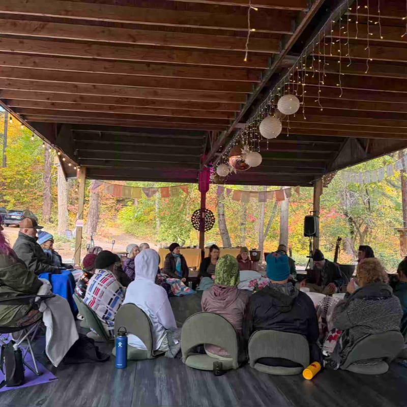 A group of people sit in chairs under a wooden pavilion decorated with string lights, facing a wooded autumn landscape.