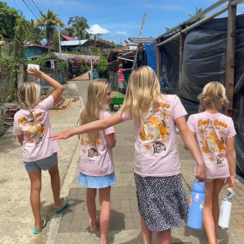 Four young girls in matching pink t-shirts walk down a sunny village street lined with rustic buildings and tropical greenery.