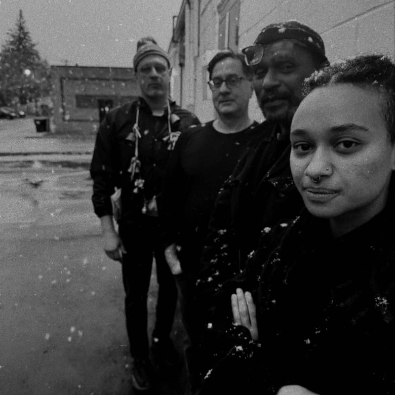 Four people stand in a line in the foreground against a backdrop of a city street and buildings during a snowfall.