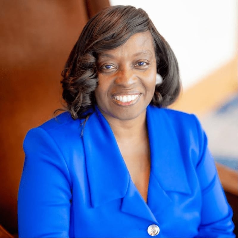 A smiling woman in a bright blue blazer sits in a brown leather chair against a blurred indoor background.
