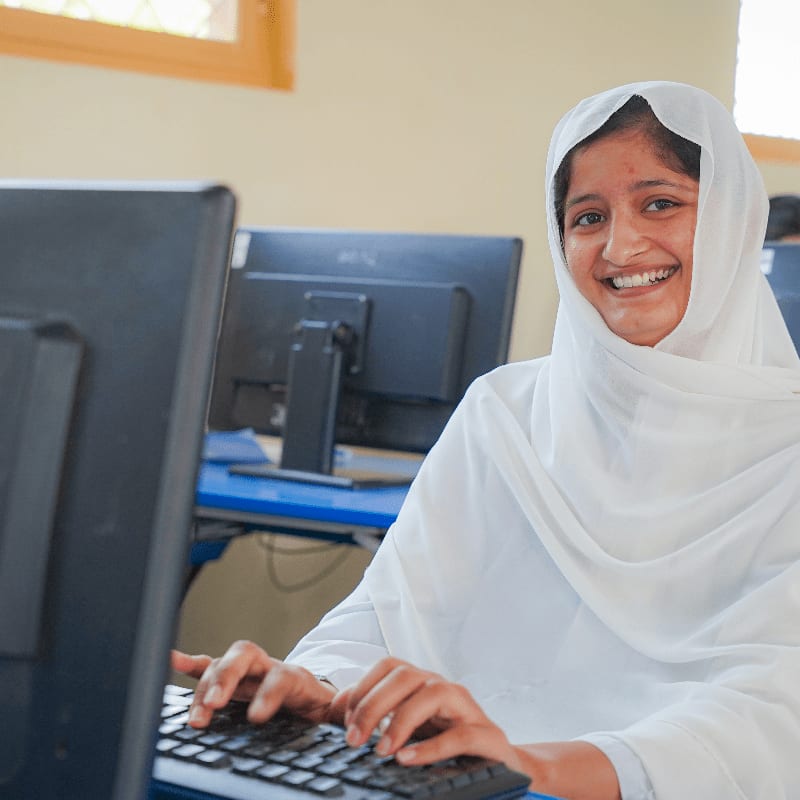 A smiling woman wearing a white hijab types on a keyboard in the foreground, with computer monitors and a classroom setting in the background.