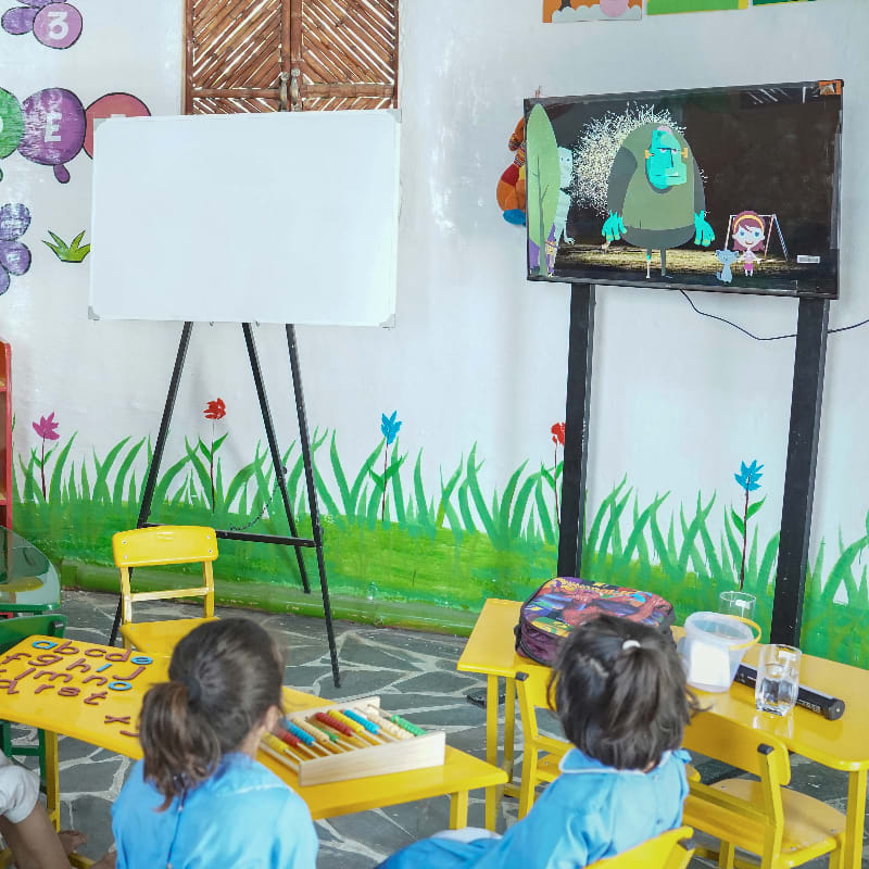 Two young children in blue smocks sit at yellow desks in a classroom, facing a white easel and a television screen displaying a cartoon.