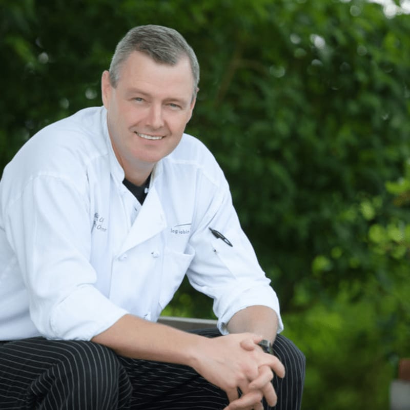 A smiling chef in a white coat and striped pants sits in the foreground against a blurred background of lush green foliage.