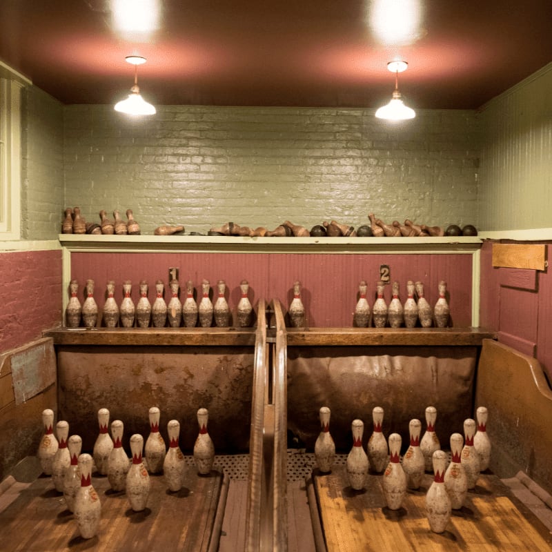 Bowling pins are arranged on wooden lanes in the foreground, with more pins lined up on shelves against a green brick wall in the background.