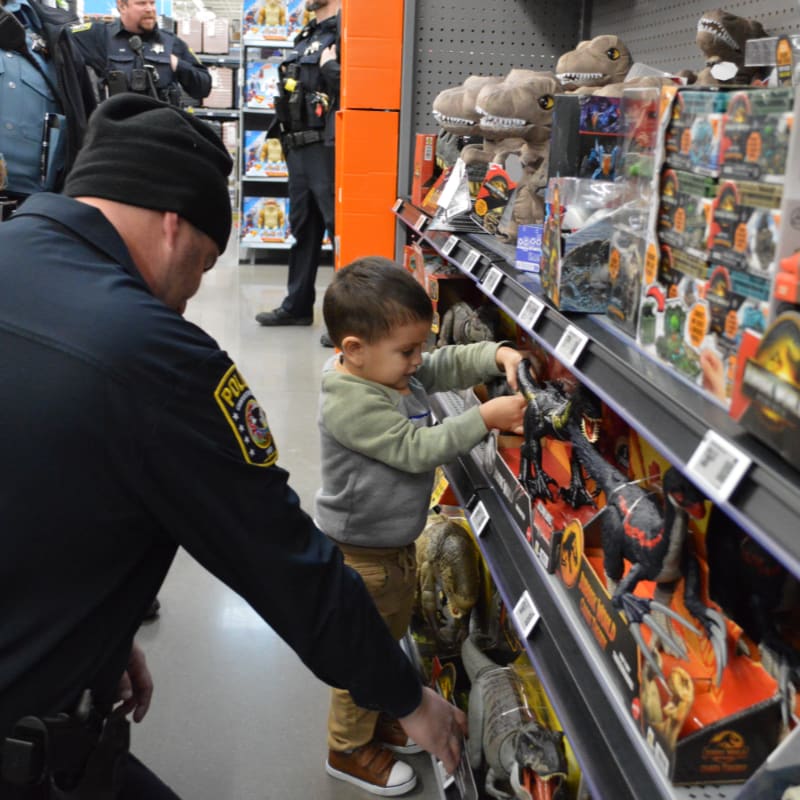 A police officer in a black beanie watches a young boy browse dinosaur toys on a store shelf, with other officers standing in the background.