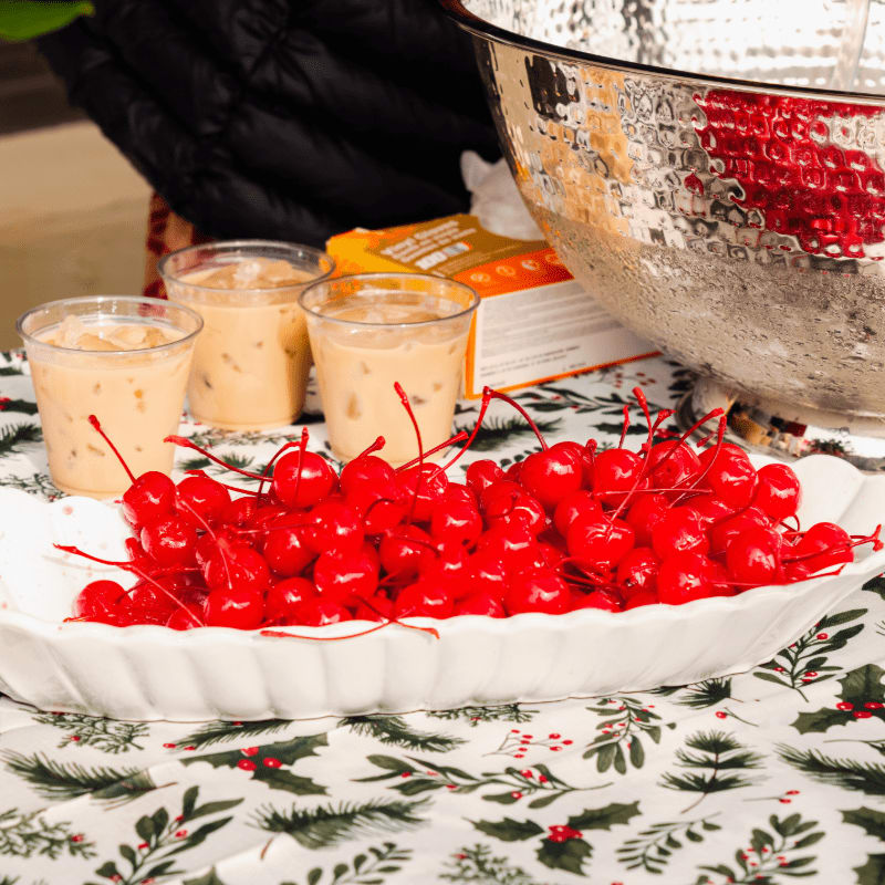 A white platter of bright red cherries sits in the foreground next to three glasses of iced coffee, with a large silver bowl and a dark jacket in the background.