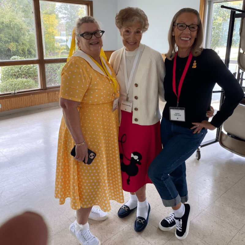 Three smiling women stand together in a brightly lit room with large windows overlooking greenery in the background.