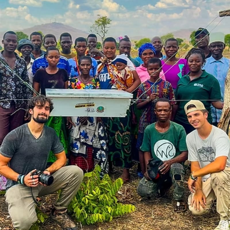 A diverse group of people gathers around a white rectangular box in a rural outdoor setting with a distant mountain under a cloudy sky.