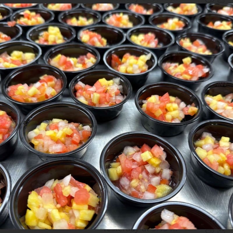 Numerous small black plastic cups filled with colorful diced salsa are arranged in rows across a metallic surface.