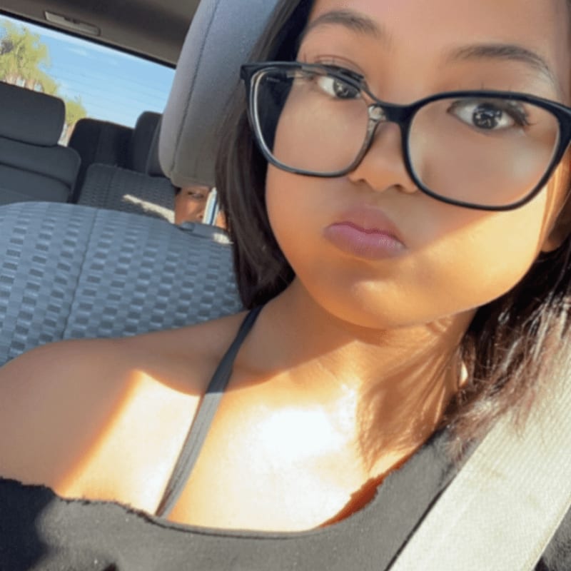 A young woman wearing black glasses and a black top is in the foreground, with the interior of a car and a bright sky visible in the background.