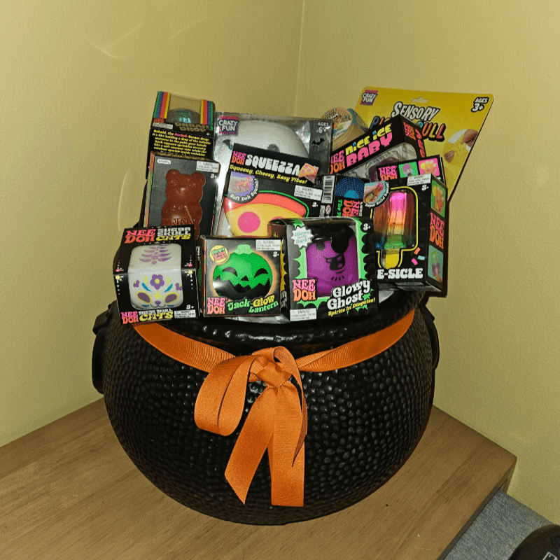 A black textured cauldron filled with various colorful Halloween-themed toys sits on a light wooden surface against a plain yellow wall.