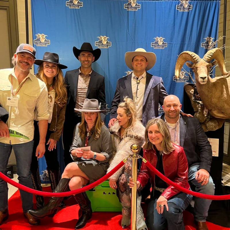 A group of people pose for a photo on a red carpet in front of a blue backdrop featuring "Wild Sheep" logos, with a large ram mount positioned to the right.