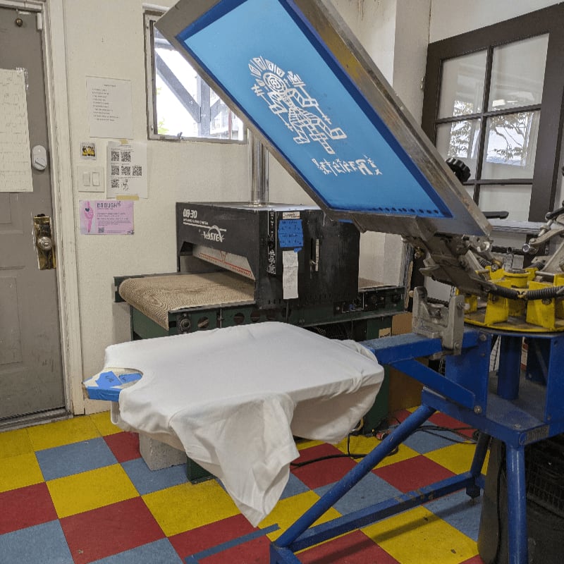 A blue screen printing press with a white t-shirt in the foreground stands before a conveyor dryer and a windowed wall in the background.