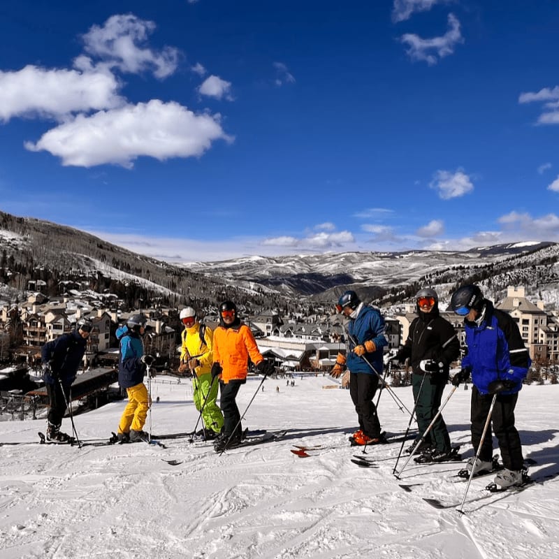 A skier in a blue suit carves through the snow in the foreground, while other skiers glide across the white slopes in the background.