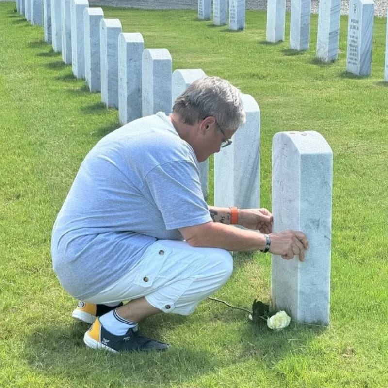 A man in a grey shirt and white shorts crouches to touch a headstone in a cemetery filled with rows of white markers.