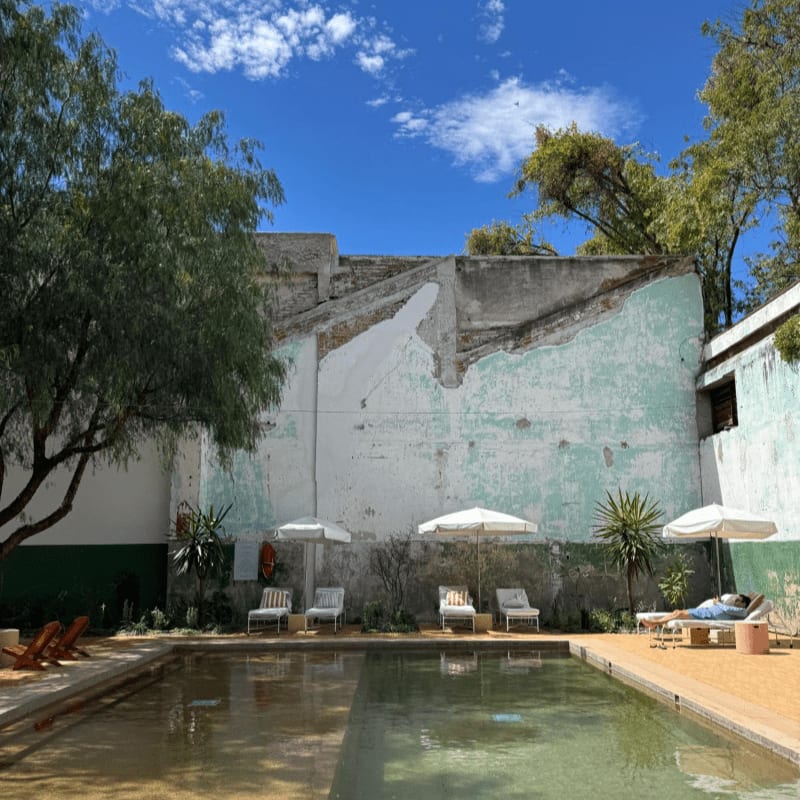 A swimming pool with lounge chairs and white umbrellas sits in the foreground against a backdrop of a weathered, light-blue wall and lush green trees under a bright blue sky.