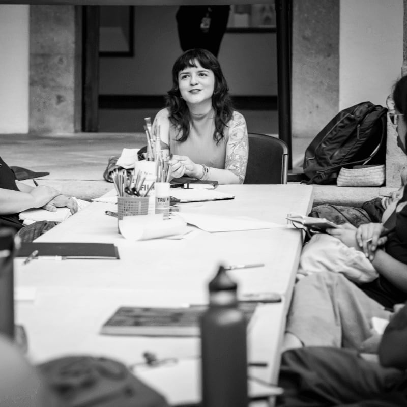 A woman sits at a white table filled with papers and pens, with a dark doorway and a backpack in the background.