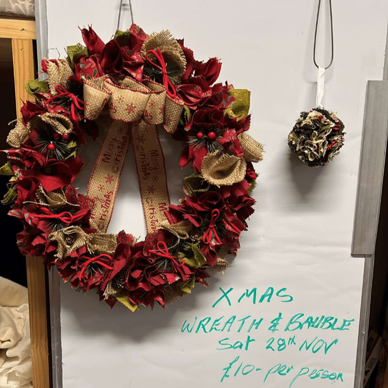 A red and burlap Christmas wreath and a small bauble hang on a white whiteboard with green handwritten text.