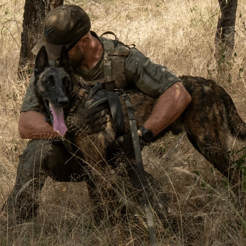 A man in military gear hugs a Belgian Malinois dog in a field of dry grass and trees.
