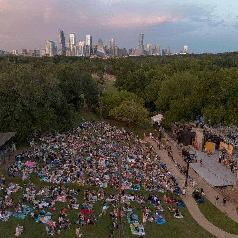 A large crowd of people lounge on blankets in a grassy field before a stage, with a dense forest and a city skyline in the background.